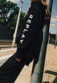 Sudadera negra con letras blancas "REPLAY" en la manga. El material parece suave, con un corte relajado y capucha. Combinada con pantalones negros a juego.