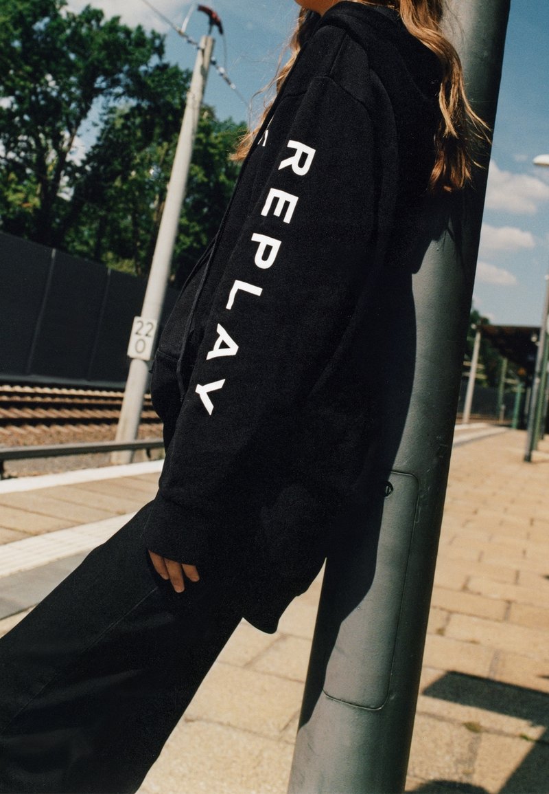 Sudadera negra con letras blancas "REPLAY" en la manga. El material parece suave, con un corte relajado y capucha. Combinada con pantalones negros a juego.