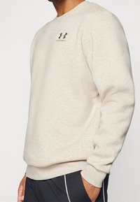 Man i beige Under Armour-tröja och svarta byxor med handen i fickan mot en enfärgad bakgrund.