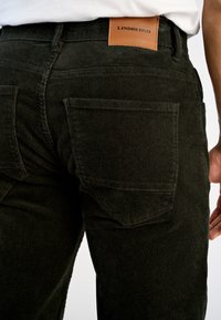 Pantaloni in corduroy verde scuro con texture a coste, dotati di due tasche posteriori e un'etichetta in pelle marrone con la scritta "LINDBERGH" in vita.