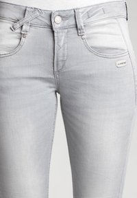 Jeans de mezclilla en gris claro con una textura suave, que cuentan con cinco bolsillos, pasadores para cinturón y un cierre de botón. Desgastado sutil en la tela.