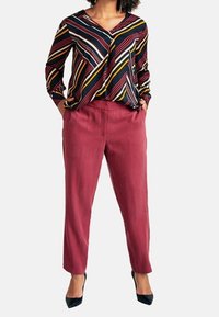 Blouse à longues manches rayée noir, rouge et jaune avec un col en V, associée à un pantalon tapered bordeaux et des escarpins noirs.