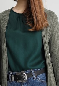 Cardigan verde a coste sopra una maglietta verde scuro, abbinato a un paio di jeans blu a vita alta. Nota cintura nera con dettagli in fibbia argentata.