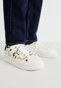 White sneakers featuring cartoon character designs, gold chain accents, and a heart detail. Textured upper with smooth rubber sole.