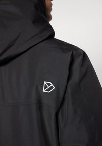 Svart jacka med huva, slät textur och en vit geometrisk logotyp på vänster axel. Enkel design utan dekorationer.