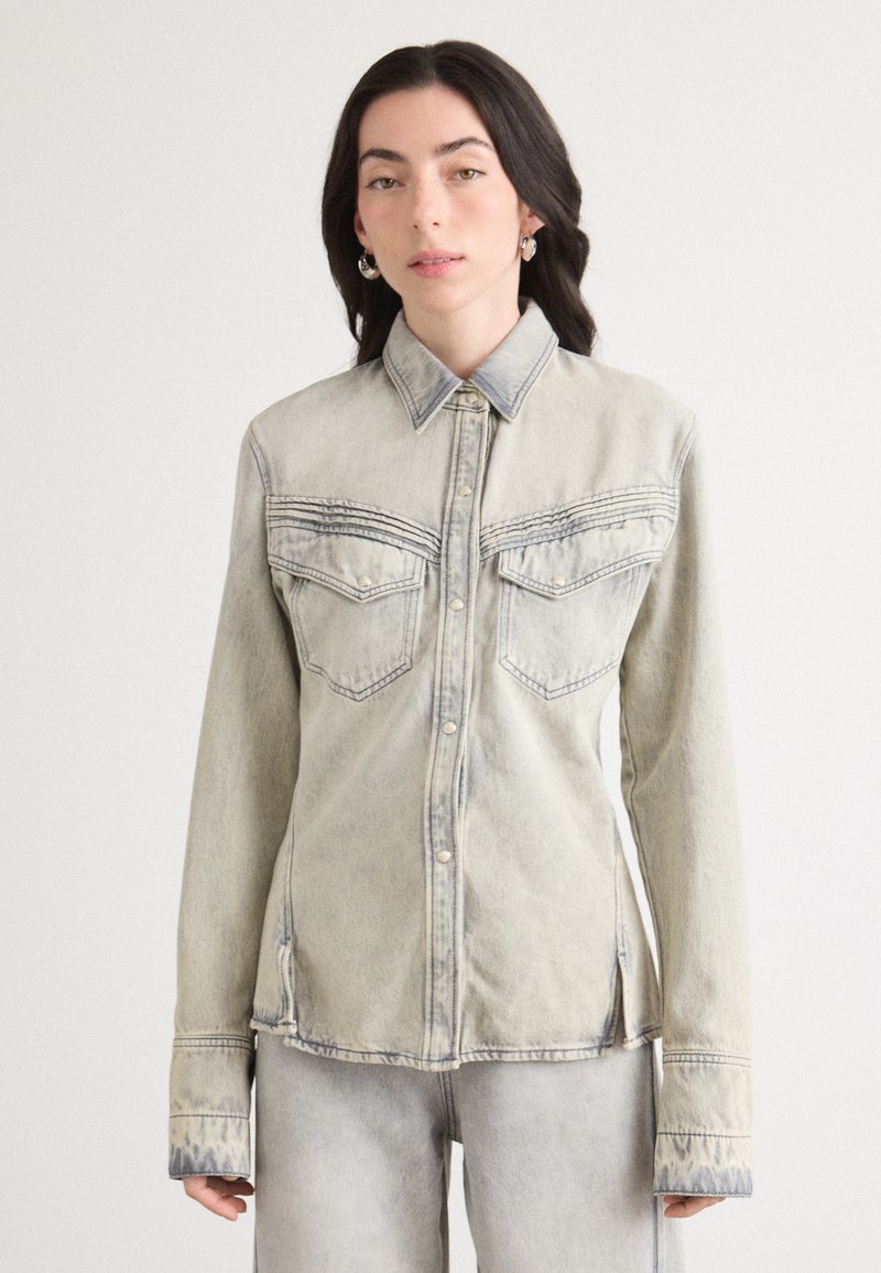Camicia di denim chiaro con bottoni frontali, due tasche sul petto e dettagli di cucitura a contrasto. Presenta un collo classico e maniche lunghe.