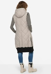 Beige gesteppte Weste mit Kapuze, knielang, kombiniert mit einem schwarzen gestrickten Saum. Gestreiftes Langarmoberteil, blaue Jeans und beige Ankle Boots.