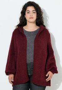 Cardigan bordeaux, lavorato a maglia, aperto sul davanti con cappuccio, maniche oversize e orlo a costine. Indossato sopra un top grigio scuro e jeans neri.
