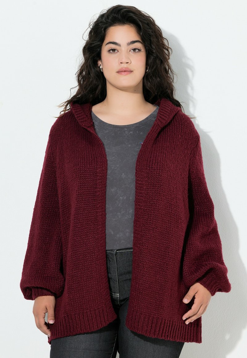 Cardigan bordeaux, lavorato a maglia, aperto sul davanti con cappuccio, maniche oversize e orlo a costine. Indossato sopra un top grigio scuro e jeans neri.