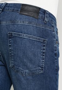 Mörkblå denimjeans med en bakficka med sydd kant och en svart läderlapp i midjan präglad med "PIER ONE."
