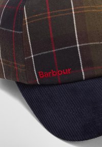 Καρό καπέλο σε σκούρο καφέ χρώμα με κόκκινες και λευκές γραμμές; διαθέτει γείσο από ναυτική κοτλέ; κόκκινο λογότυπο "Barbour" ραμμένο στο πλάι.