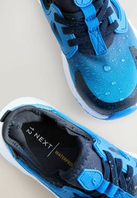 Baskets étanches bleues et noires avec une surface texturée, des lacets et un marquage distinctif. Gouttes d'eau visibles sur le matériau supérieur.