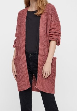 Femme portant un cardigan en tricot épais couleur vieux rose avec les manches retroussées, des poches devant, un chemisier noir et un pantalon noir taille haute.