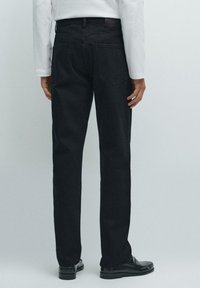 Jeans in denim nero con taglio dritto, cinque tasche e dettagli di cucitura discreti, indossati con scarpe nere e una camicia bianca a maniche lunghe.