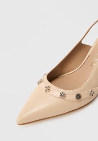 Zapato beige de punta puntiaguda con tira trasera y una correa decorativa con pequeños adornos metálicos florales y geométricos.