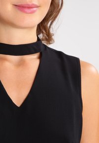 Schwarzes Kleid mit V-Ausschnitt und einem Stoff-Choker-Detail. Das Material wirkt glatt mit einer leicht strukturierten Oberfläche. Ärmelloses Design.