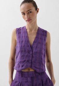 Vesta fără mâneci, de culoare purpurie, cu guler tip notch, având două nasturi îmbrăcați în țesătură, realizată dintr-un material texturat și ușor. Accentuată de un model în carouri.