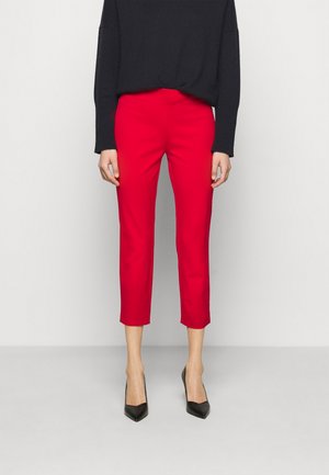 Persona con pantalones cortos de color rojo brillante, top negro de manga larga y tacones altos negros de punta puntiaguda, de pie contra un fondo liso.