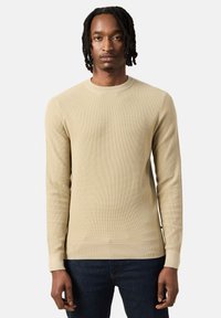 Maglione a maniche lunghe a costine beige con scollo a girocollo e polsini aderenti, abbinato a jeans blu scuro. La texture sembra morbida e lavorata.
