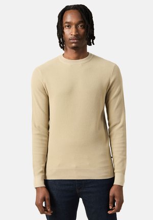 Beige geribbelde gebreide lange mouwen trui met een ronde hals en aansluitende manchetten, gecombineerd met donkerblauwe jeans. De textuur lijkt zacht en gestructureerd.