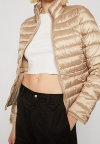 Helle beige Daunenjacke mit horizontaler Steppung, hohem Kragen und Reißverschluss. Kombiniert mit einem weißen Crop-Top und schwarzen Hosen.