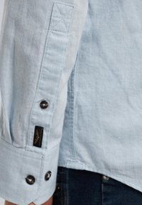 Manga de camisa de lino azul claro con un puño abotonado que presenta dos botones oscuros, costura decorativa y una pequeña etiqueta de marca negra.