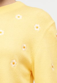 Pull tricoté jaune avec de petites marguerites blanches et orange brodées éparpillées sur l'épaule et la manche.