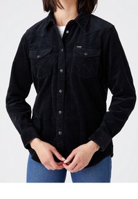 Black corduroy shirt with a button-up front, two chest pockets, and a textured fabric. Features a pointed collar and long sleeves.