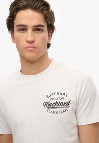 Witte katoenen t-shirt met ronde hals, voorzien van een zwarte geprinte logo met de tekst "SUPERDRY PRECISION Machined COPPER LABEL" op de borst.