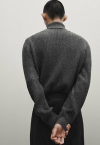 Szary sweter z prążkowanej dzianiny z wysokim kołnierzem, noszony z dłońmi splecionymi za plecami. Dopasowany krój, fakturowany materiał. Minimalistyczny design, neutralne odcienie.