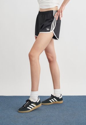 Persoană purtând pantaloni scurți negri Adidas și adidași negri Adidas cu dungi albe, stând pe covor albastru pe fundal alb.