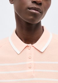 Κοντινό πλάνο ενός ατόμου που φοράει ένα ροζ πλεκτό πόλο με λευκές ρίγες και τρία κουμπιά, εμφανίζοντας τον λαιμό και το κάτω μέρος του προσώπου.