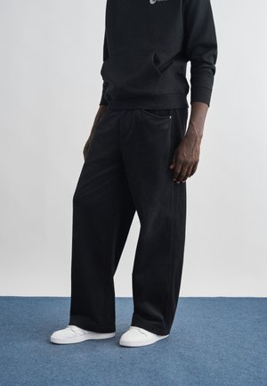 Isik, kes kannab musti laiapõlve sametpükse, musta dressipluusi ja valgeid tossusid, seisab sinisel vaibal halli tausta ees.