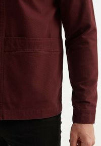 Veste bourgogne en tissu texturé, avec une poche avant et une coupe droite, montrée de la taille vers le haut sur fond noir.