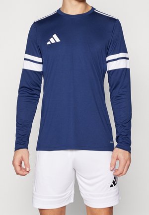 Mężczyzna ubrany w granatową sportową koszulkę z długim rękawem z białymi paskami oraz białe krótkie spodenki sportowe z czarnym logo.