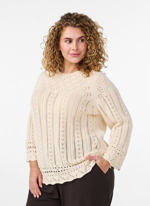Vrouw met krullend haar die een crèmekleurige gebreide trui met kantpatronen draagt, staat met één hand in de zak van een donkere broek en kijkt opzij.
