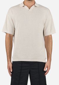 Hombre vistiendo una camiseta de polo de punto beige de manga corta con un patrón texturizado y pantalones negros, de pie contra un fondo liso.