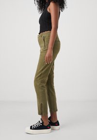 Olive green, tapered cargo pants with side zipper pockets and distressed texture, paired with black sneakers featuring red cherry accents.