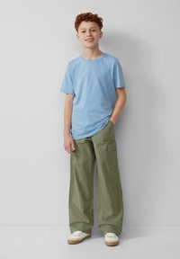 Lichtblauwe katoenen t-shirt, losse olijfgroene cargo broek met zijzakken en witte sneakers met beige accenten; staande pose tegen een neutrale achtergrond.