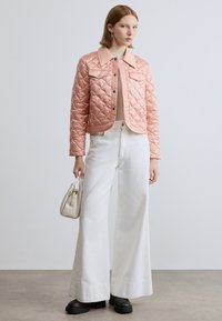 Ροζ μπουφάν με καπιτονέ υφή και γιακά, συνδυασμένο με κρεμ πουλόβερ και φαρδιά λευκά παντελόνια. Το μοντέλο κρατά μια μικρή μπεζ τσάντα.