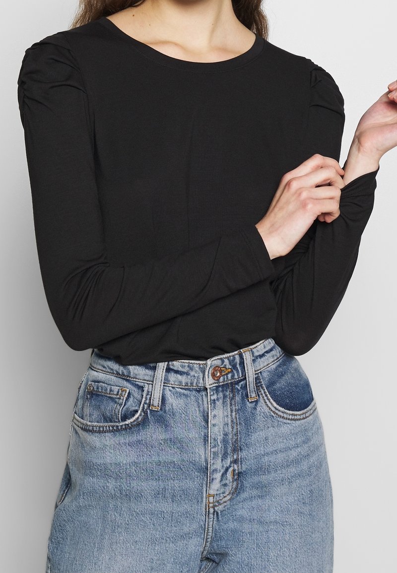 Top noir à manches longues avec détail froncé aux épaules, en tissu lisse. Associé à un jean en denim bleu clair taille haute.