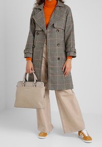 Karo-Trenchcoat mit breiten Revers, braunen Knöpfen und langem Schnitt. Beiger Handtasche aus Kunstleder und beigefarbenen, weit geschnittenen Hosen. Gelbe Sneakers mit weißen Sohlen.