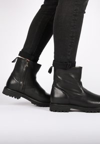 Botines de cuero negro con un acabado suave, cremallera lateral y suela de goma. Diseñados con tiradores en el cuello para facilitar su colocación.