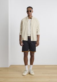 Modell iført en lys beige skjorte med knapper, hvit t-skjorte, svarte shorts med brune mansjetter, hvite sokker og beige joggesko. Brun belte.