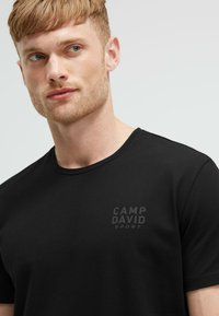 Black short-sleeve t-shirt with a crew neckline, featuring a textured fabric and a subtle "CAMP DAVID SPORT" logo on the chest.