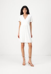 White, short-sleeved dress with a wrap design, featuring a textured floral pattern. Paired with strappy, light tan high-heeled sandals.
