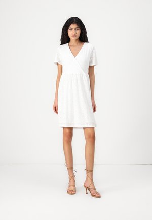 Robe blanche à manches courtes avec un design croisé, présentant un motif floral texturé. Associée à des sandales à talons hauts légères et beige.