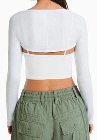 Kvinna iklädd en vit långärmad crop top över en vit spaghetti strap-top, tillsammans med högmidjade olivgröna cargobyxor.