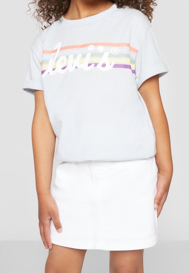 Dziecko ubranie w jasnoszarą koszulkę Levi's z pastelowymi paskami w kolorze tęczy i białą spódnicę, stojące na tle jednolitego tła.