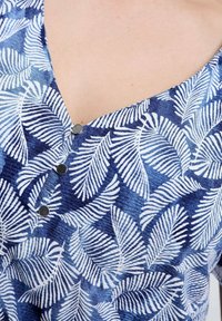 Blauw-wit stoftop met bladmotief, een V-hals en drie grijze knopen in het midden. Het materiaal lijkt licht en met textuur.